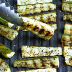 tongs holding pieces of grilled zucchini on grill pan