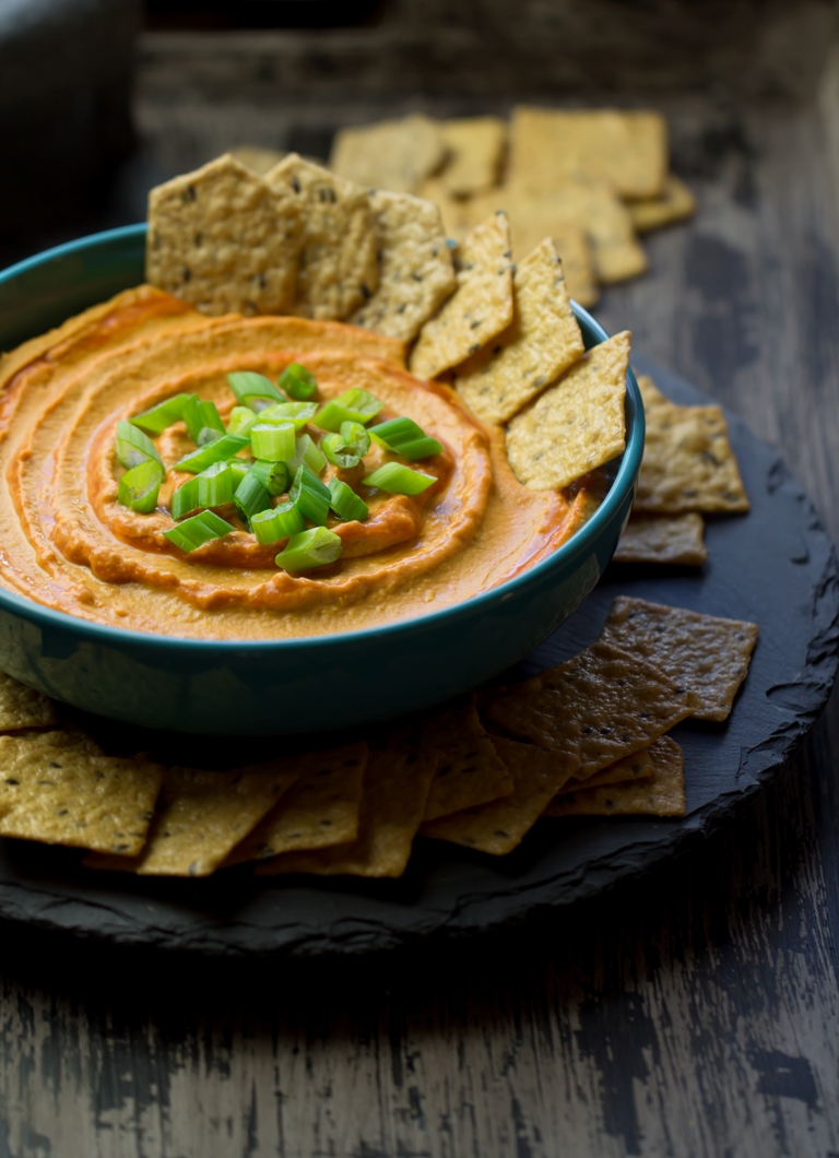 Bowl of spicy vegan buffalo hummus with crackers and green onions on a platter.