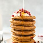 stack of vegan gingerbread pancakes with whipped cream and pomegranate perils and syrup drizzling